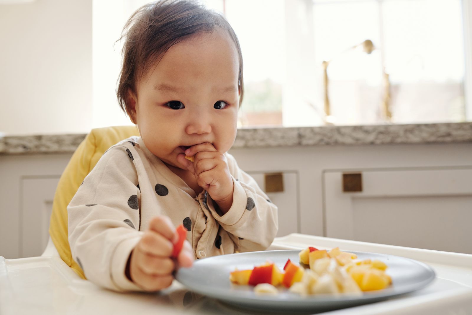 A high-chair tray with a small spread of first foods laid out: avocado wedge, banana spear, mashed beans on toast, steamed broccoli, meat strip