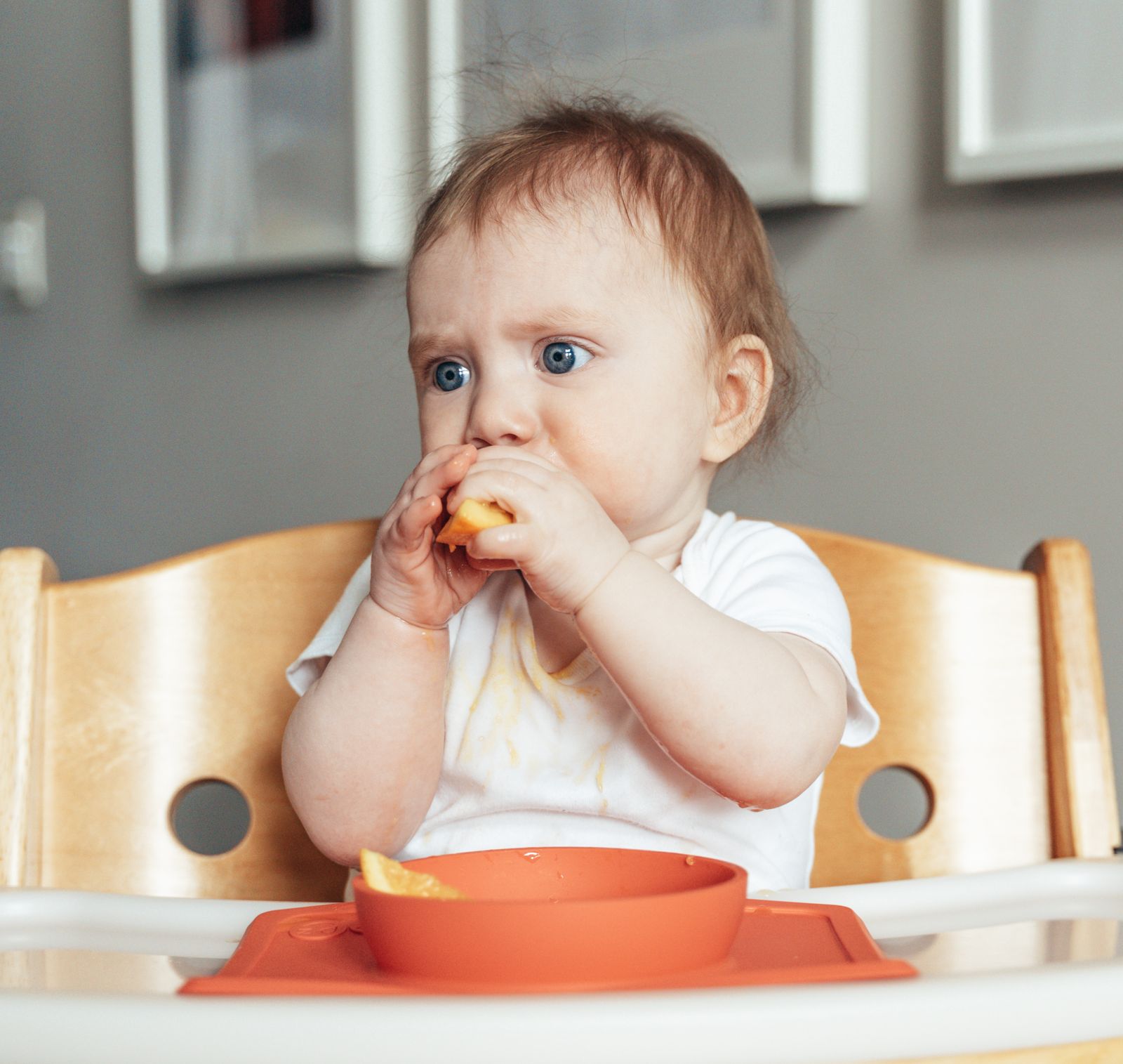 A baby in a high chair reaching for a strip of banana, hand raised toward mouth, focused expression