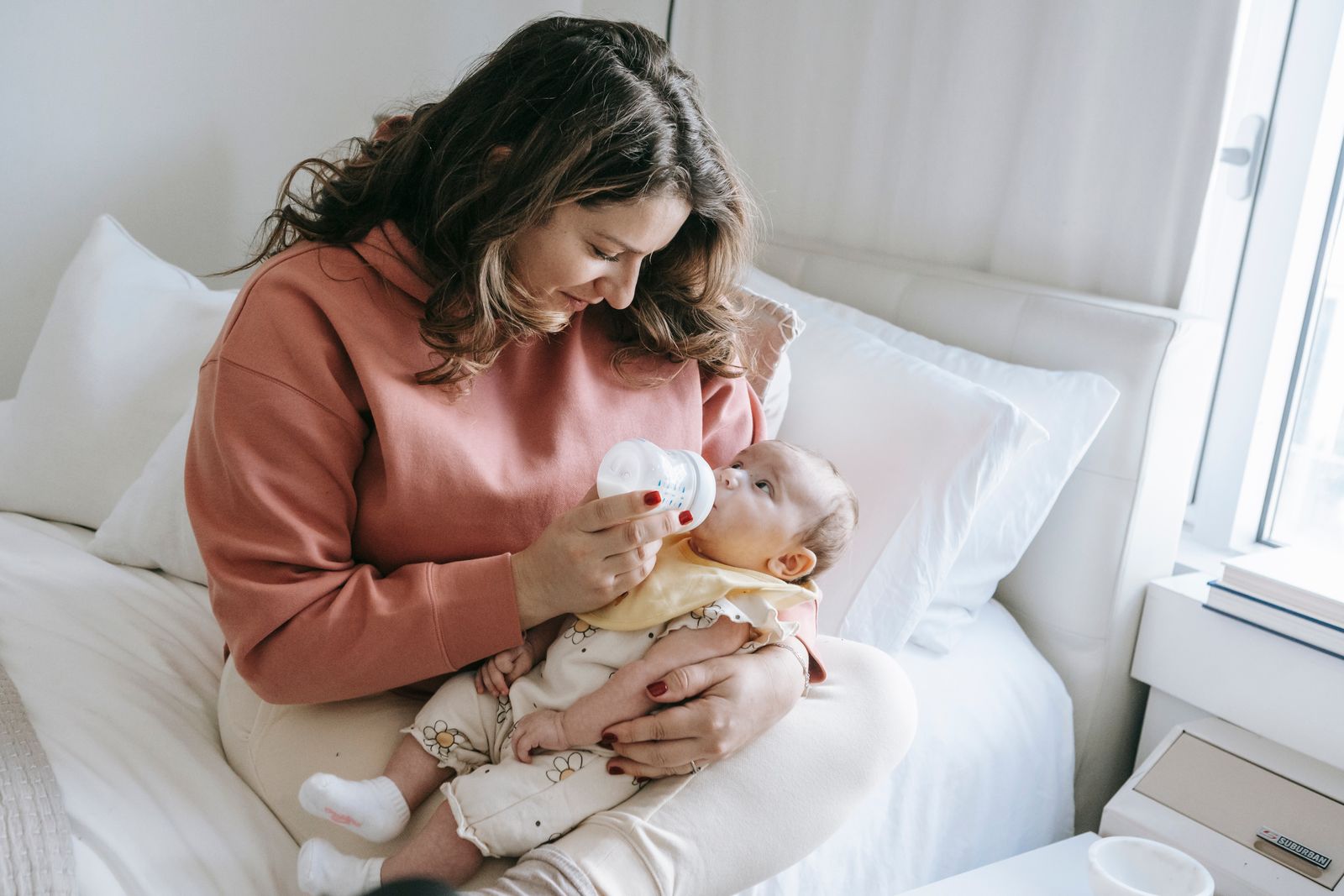 Mother in a rust sweater feeding her baby from a bottle in a semi-upright hold on a bed