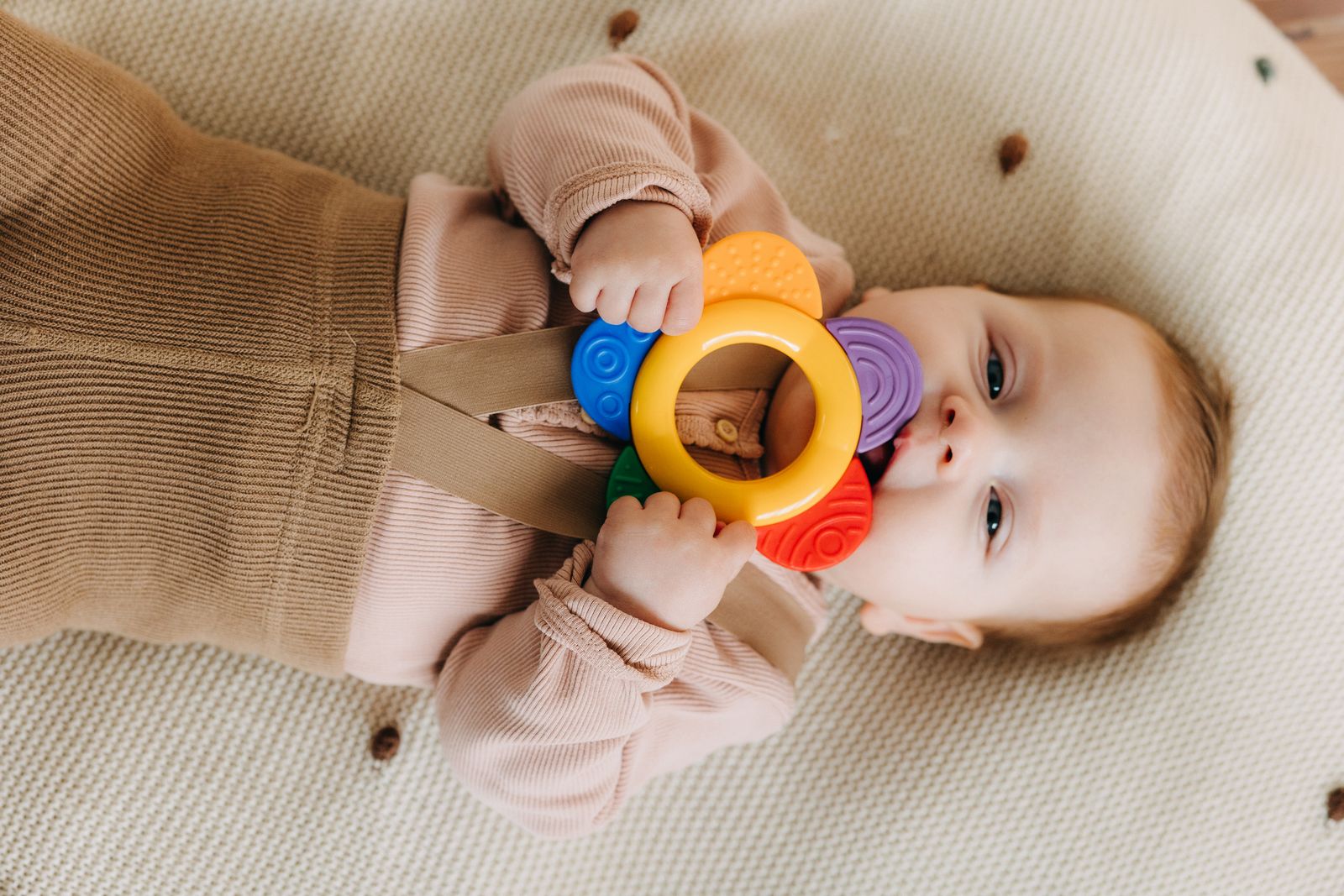 A baby lying down chewing on a colorful silicone teething ring