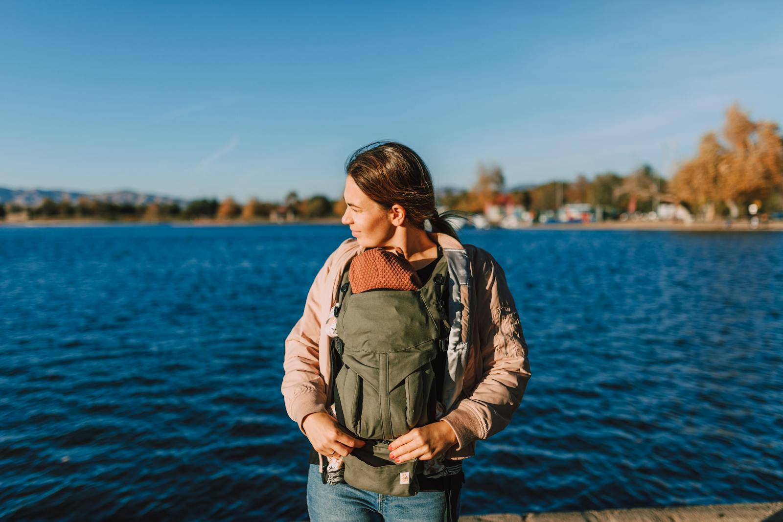 mom smiling outdoors holding baby in a soft carrier near water