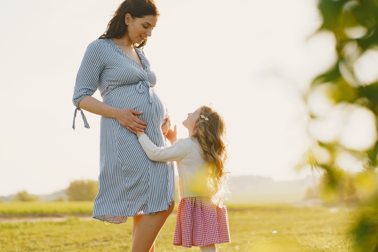A pregnant mom in a striped dress standing in a sunny field while her young daughter looks up at her belly