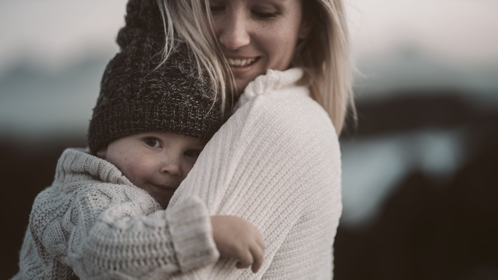 A mom in a cream sweater holding her cozy, knit-capped toddler close and smiling