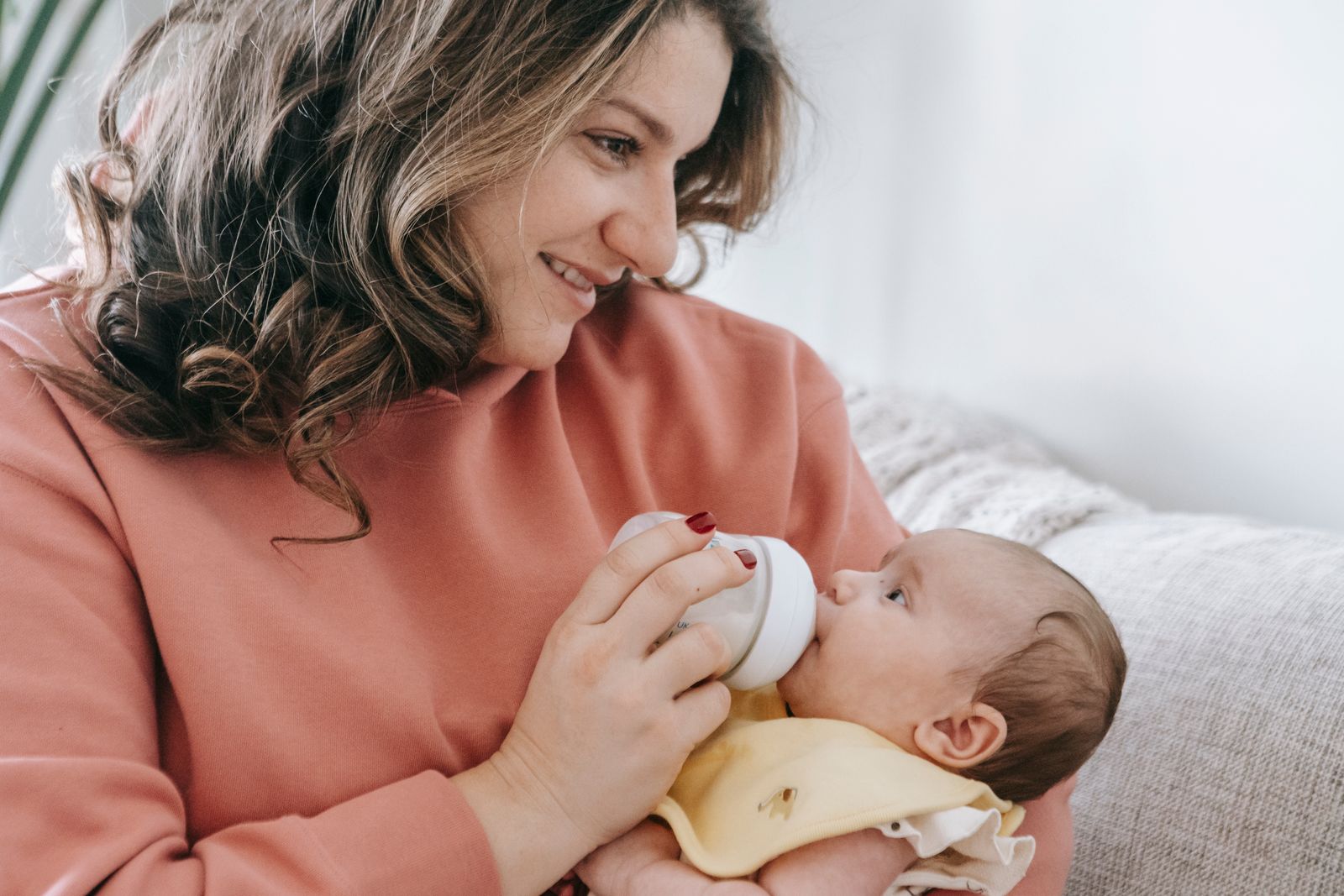 A mother in a coral sweater bottle feeding her baby on the couch