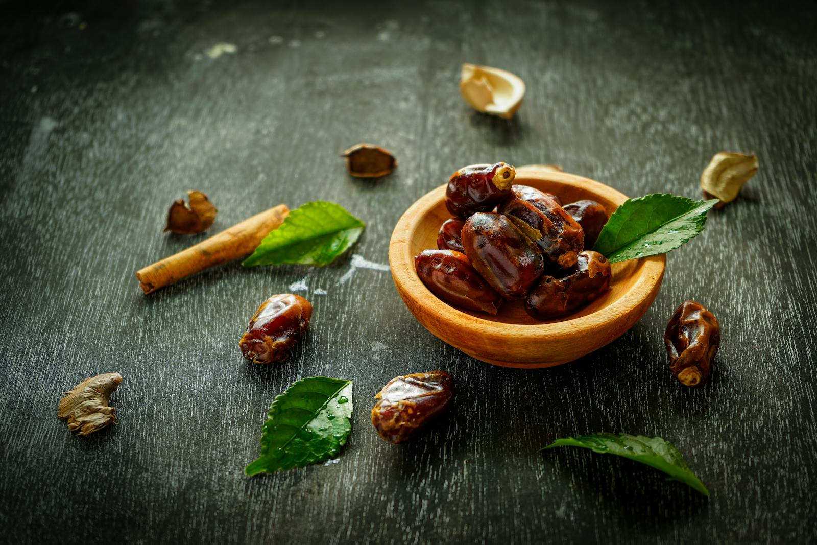 Fresh dates in a small wooden bowl on a dark wood surface with cinnamon and green leaves arranged around it.
