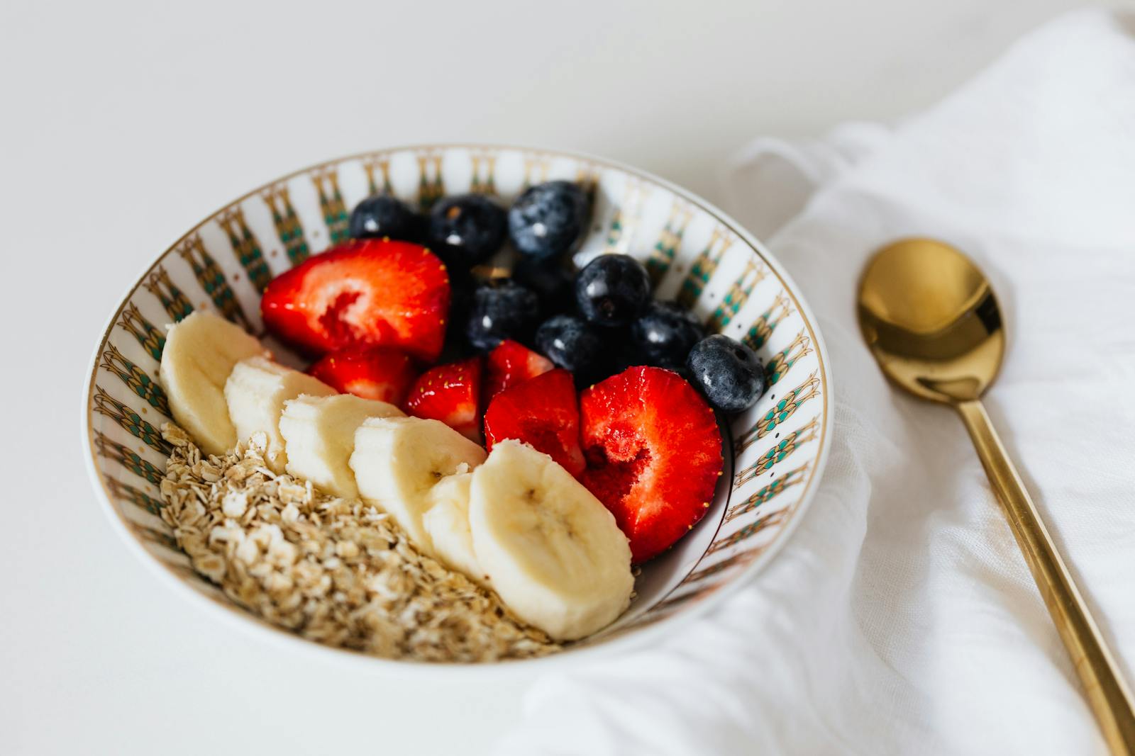 A bowl of oats with sliced bananas, strawberries, and blueberries on a white surface, placed on a folded white cloth with a gold spoon nearby.