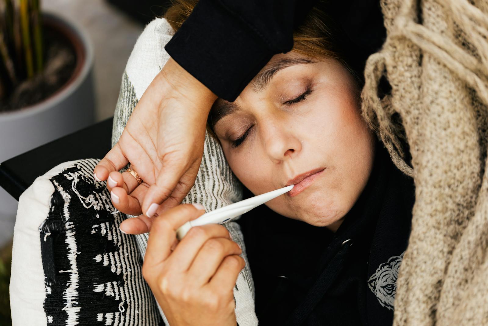 Woman lying on couch checking thermometer wrapped in cozy blanket