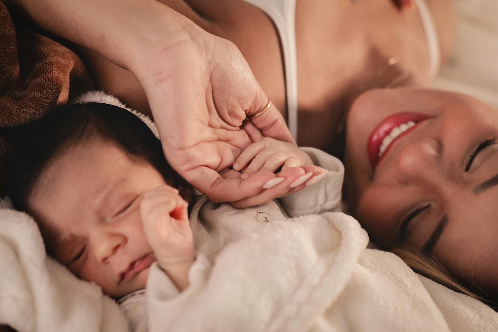 Mother and newborn holding hands while lying together, fingers intertwined in a quiet tender moment