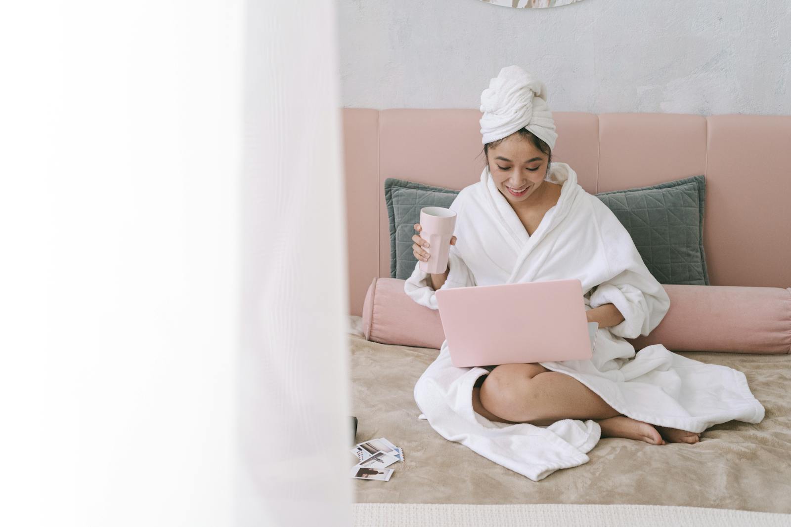Mom in a bathrobe sitting on a bed with coffee, looking thoughtfully at a laptop in a soft pink and white bedroom