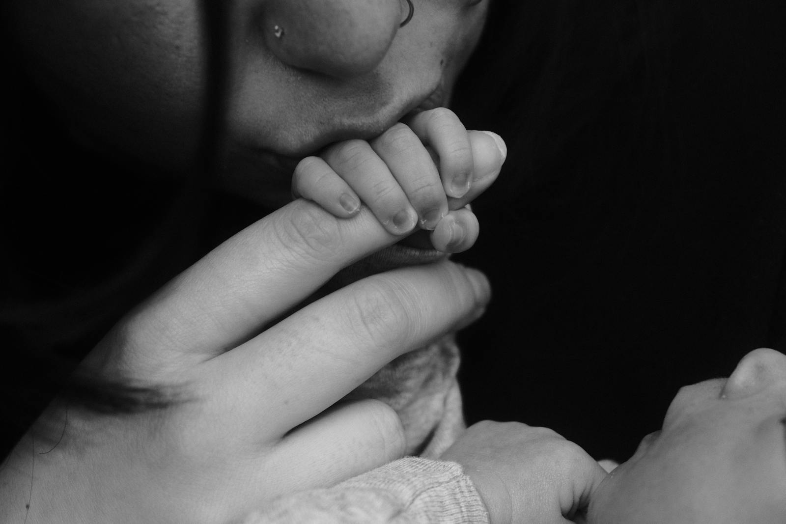 Close-up black and white photo of a baby's tiny hand wrapping around an adult's finger, with the parent kissing the hand