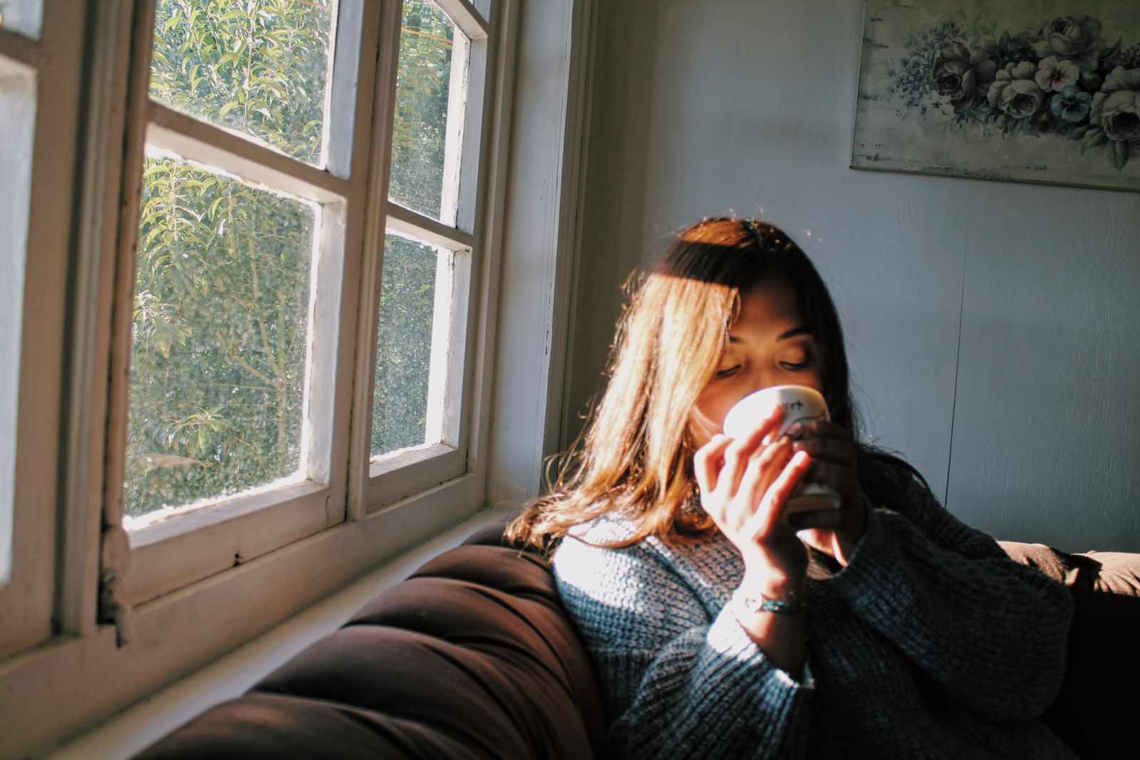A woman wrapped in a sweater resting by a sunlit window with a warm drink