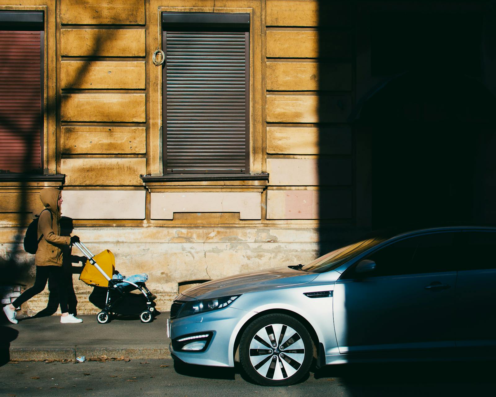 A mother walks with a baby stroller along a sunlit sidewalk in the morning