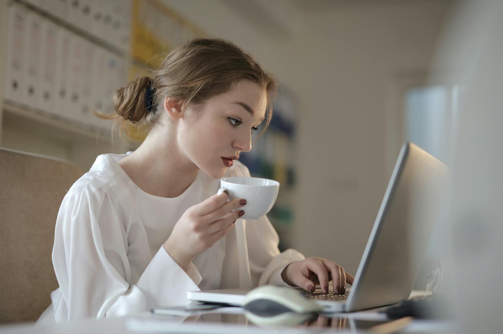 A focused woman in a white blouse working at a laptop with a coffee cup in hand