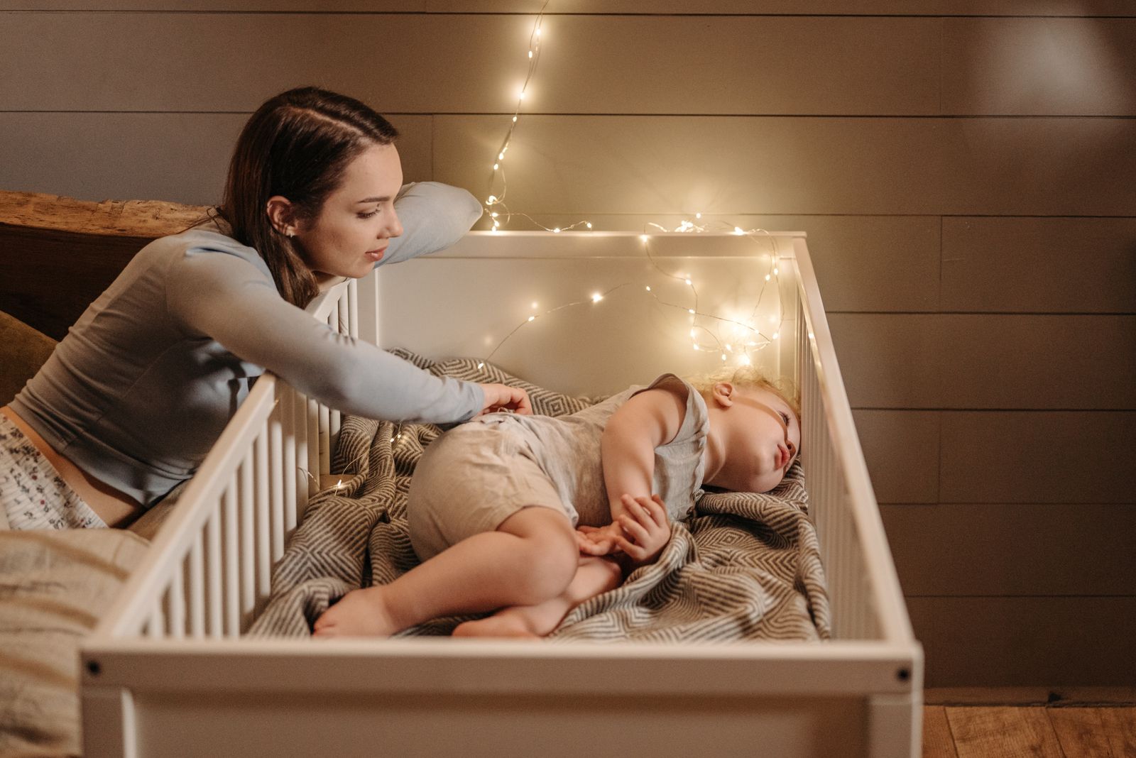 A parent leans over the crib to gently settle a sleepy baby under warm lights