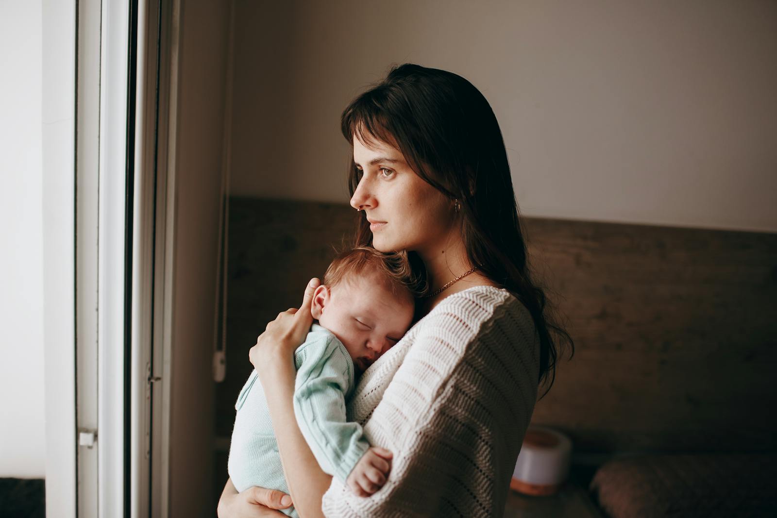 A mother standing by a window holding her baby, looking calm and a little tired
