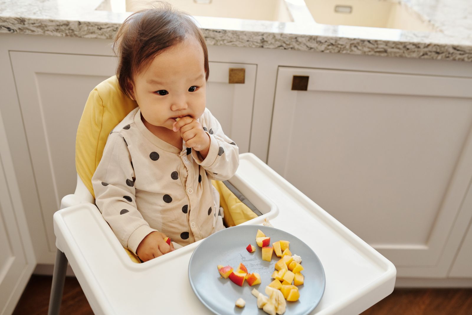 A baby sitting up in a high chair bringing a piece of soft food to their mouth on their own