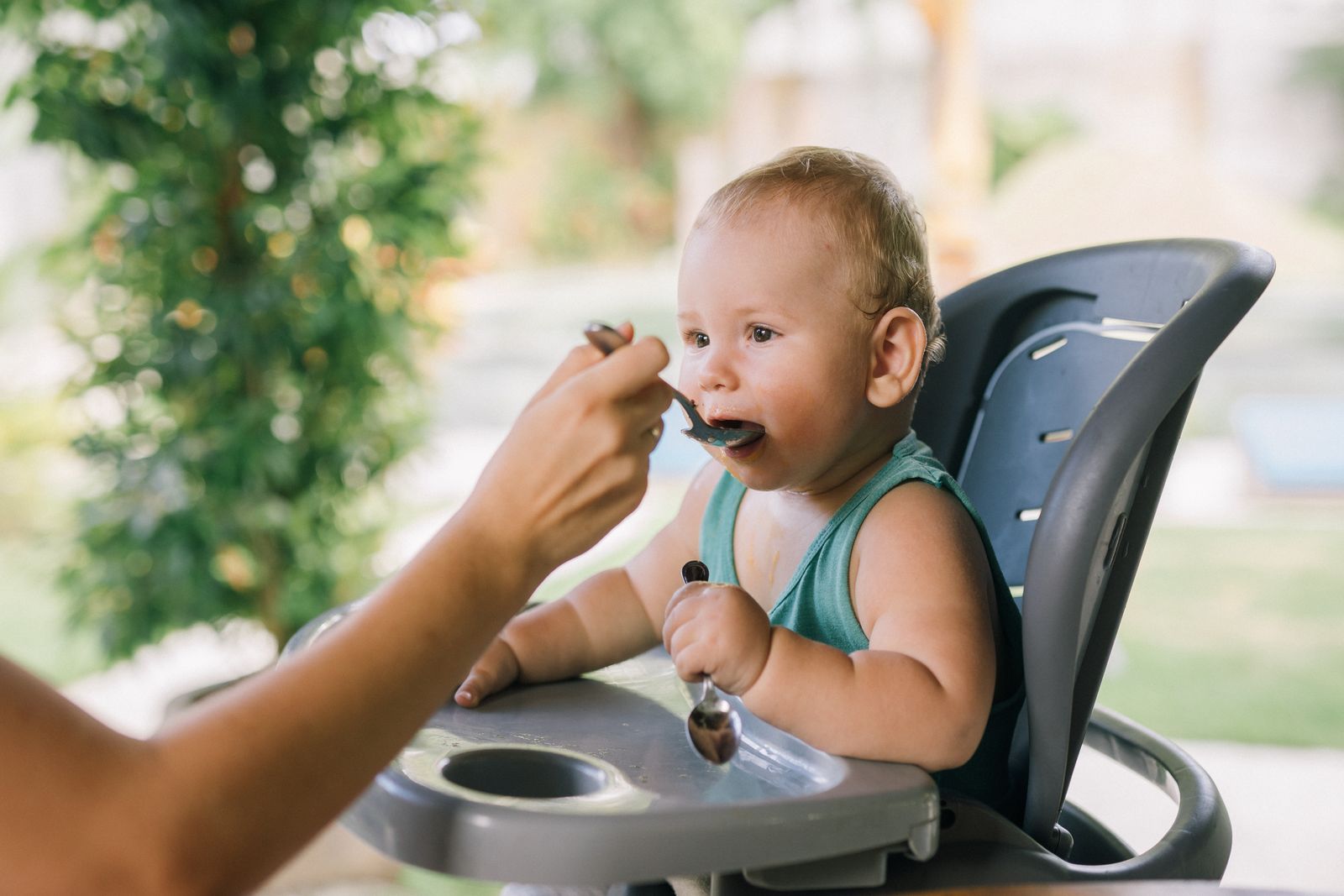 A parent spoon-feeding a smooth puree to a baby seated outdoors in a high chair