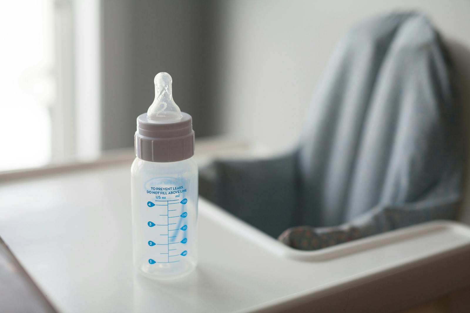 A clean baby bottle sitting on a white surface near a soft chair