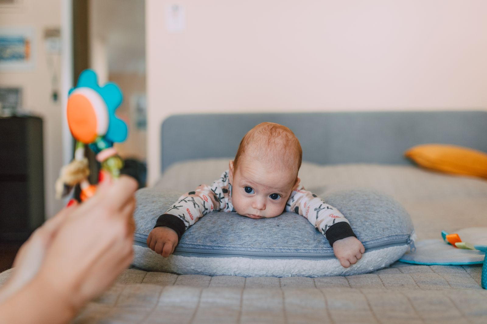Baby on tummy over a small pillow, looking up at a colorful toy held by a parent