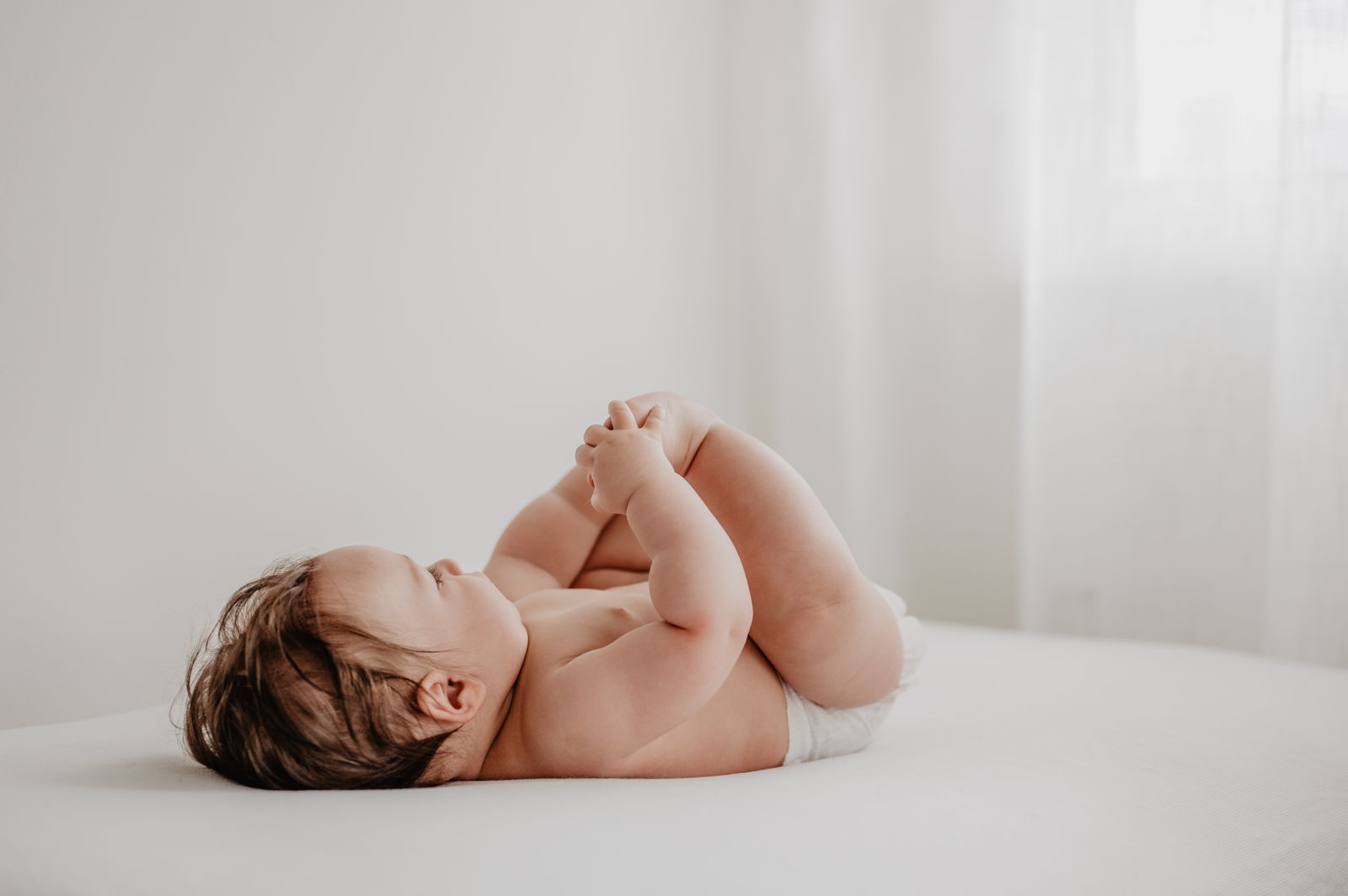 A baby resting in soft natural window light on a white bed