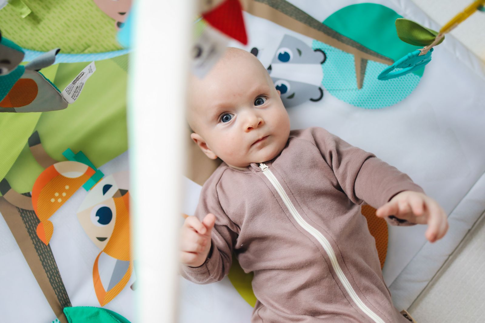 A baby sitting on a soft mat, playing with a small teether, alert and content, morning light on a window sill in the background