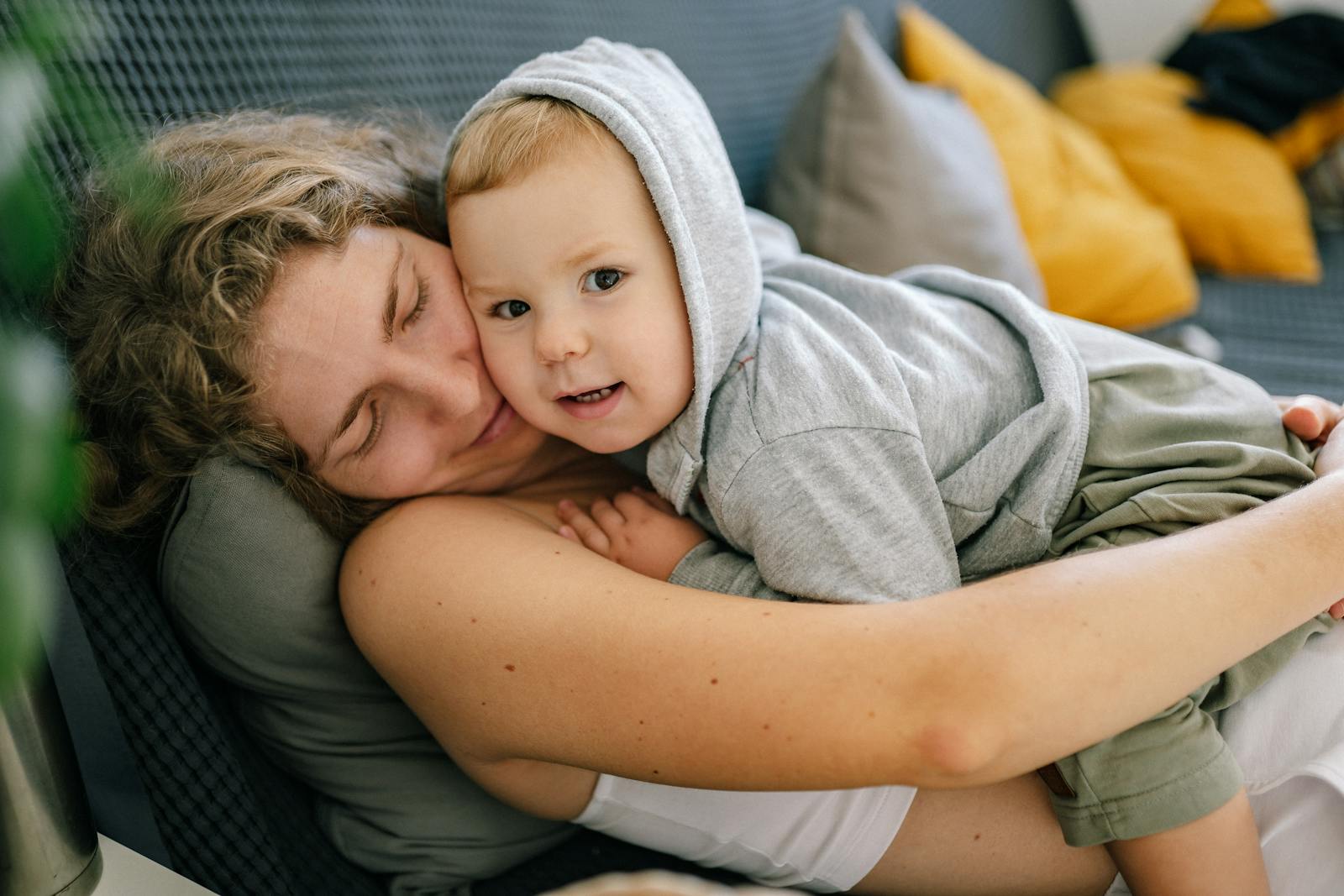 A mother cuddling her toddler close on a sofa at home, both relaxed and content