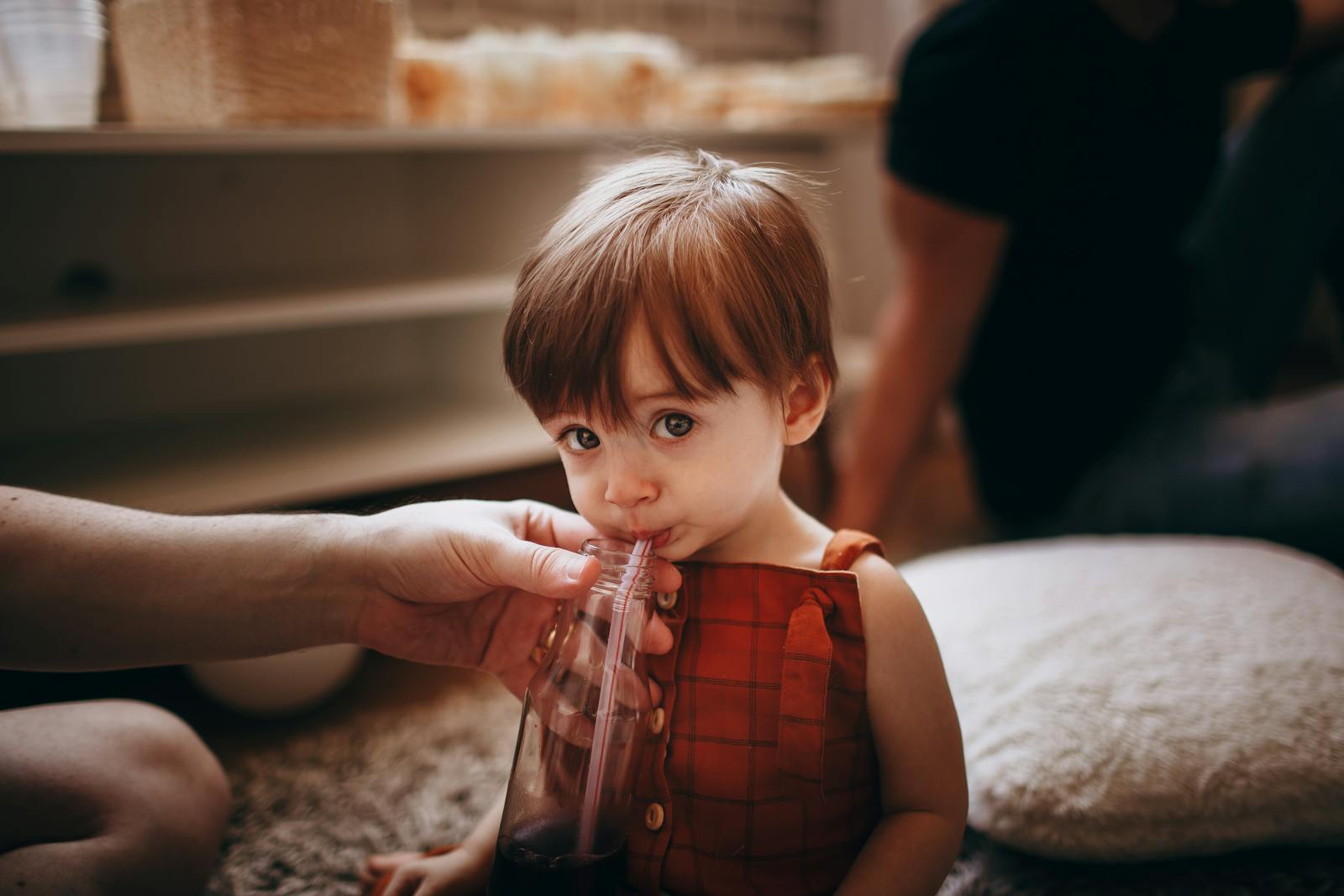 A toddler in a red outfit drinking from a cup with a straw held by a parent